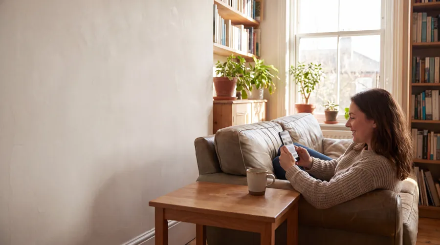 A woman relaxed on a sofa in a sunlit living room reading news on her phone, with open wall space on the left
